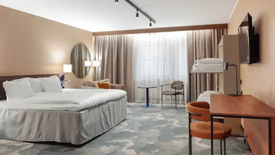 Et pænt hotelværelse med en stor seng, sofa og skrivebord har varm belysning og beige vægge. Et vindue med tunge gardiner oplyser rummet, inklusive et vægmonteret TV.