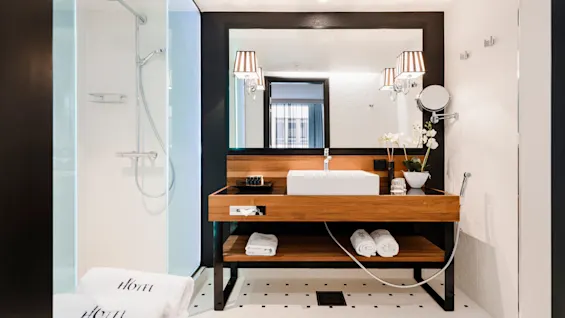 Moderne badeværelse med en håndvask og spejl indrammet i træ ved siden af en glasbrusekabine. Hvide håndklæder med teksten "HOTEL". Elegant belysning og orkidédekoration.