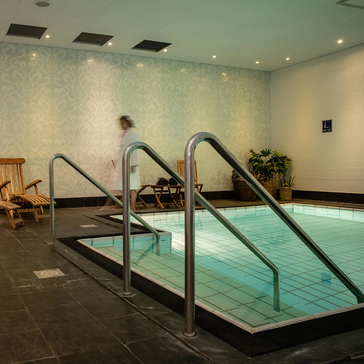 Pool omgivet av bruna trästolar i ett spa med gröna växter och en person som går förbi. Mjuk belysning skapar en avkopplande atmosfär. Grå handdukar hänger på väggkrokar. 