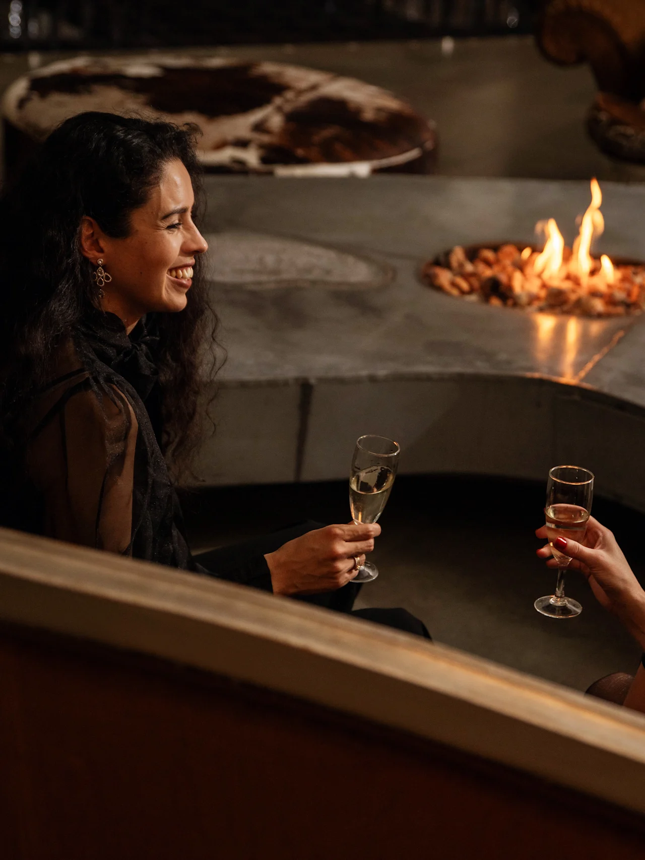 Two people sit by a circular fire pit, holding champagne glasses, in a cozy lounge with plush seating and warm lighting.