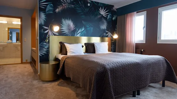 A double bed with brown and gold bedding rests against a vibrant tropical-patterned wallpaper. Nightstands with lamps flank the bed. Two windows on the right provide natural light.
