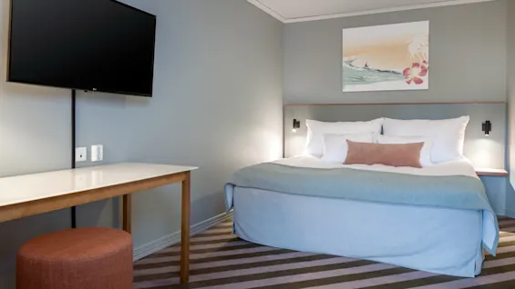 A bed with white and blue bedding is positioned in a hotel room. A painting hangs above the bed. A table and stool sit opposite, with a flat-screen TV on the wall.