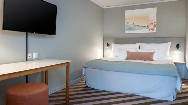 En seng med hvitt og blått sengetøy er plassert i et hotellrom. Et maleri henger over sengen. Et bord og en krakk står overfor, med en flatskjerm-TV på veggen.