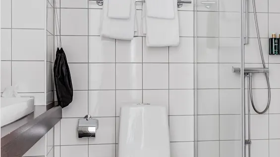 Toilet stands in a modern, tiled bathroom with a nearby shower; walls are white, and the floor is dark tile. Towels hang above, and a black bag hangs from a hook.