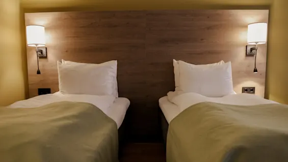 Two twin beds with white pillows and green blankets sit side by side, flanked by wooden headboards and wall-mounted lights in a softly lit room.