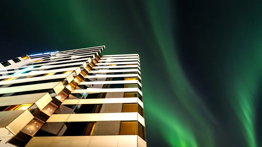 En modern hotellfasad upplyst på natten, med livfulla gröna norrsken som dansar över den mörka himlen ovanför.