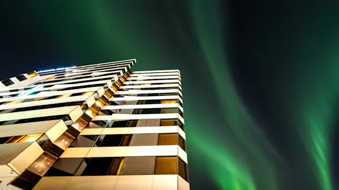 A modern hotel facade illuminated at night, with vibrant green northern lights dancing across the dark sky above.