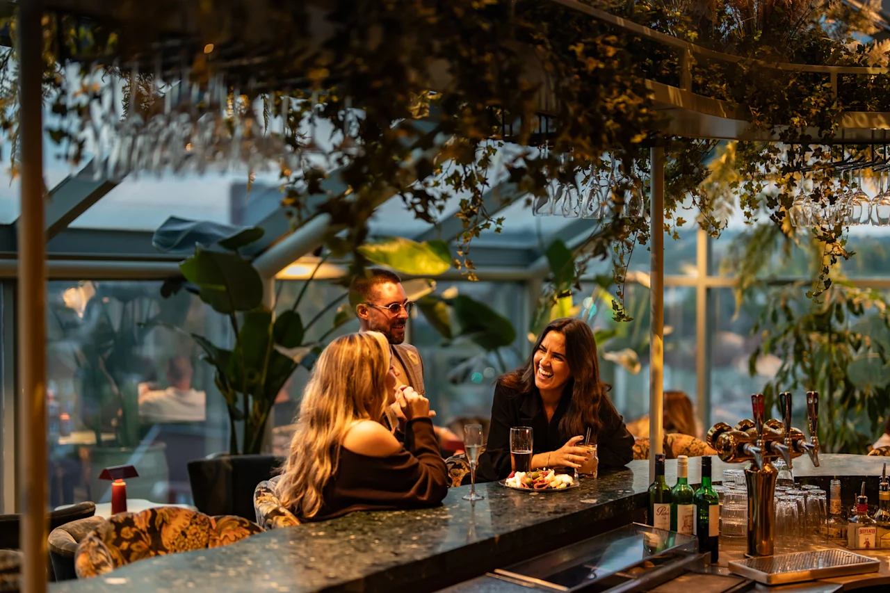 Ihmiset nauttivat juomista ja keskustelusta vilkkaassa baarissa, jossa on rehevää kasvillisuutta ja valoisa, avoin tunnelma.