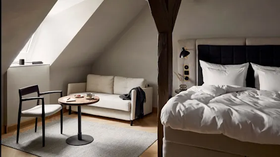 A cozy bedroom with a bed under a slanted ceiling, adjacent to a sofa and wooden table with a chair on a gray rug.