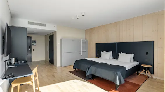 Et lyst og moderne hotelværelse med to enkeltsenge, et skrivebordsområde og lyse trædetaljer, der tilbyder et behageligt ophold.