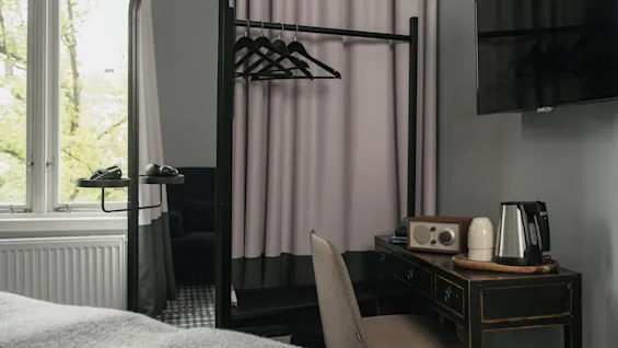 Ett litet hotellrum med ett skrivbord och en stol, en vattenkokare och en vintage radio. En TV är monterad på väggen. En garderob med galgar finns i närheten.