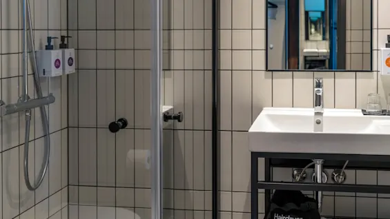 Dusj med metallarmatur og såpedispensere i flislagt bad. Ved siden av er en vask under et speil som reflekterer flere bilder. Tekst inkluderer "Hairdryer."