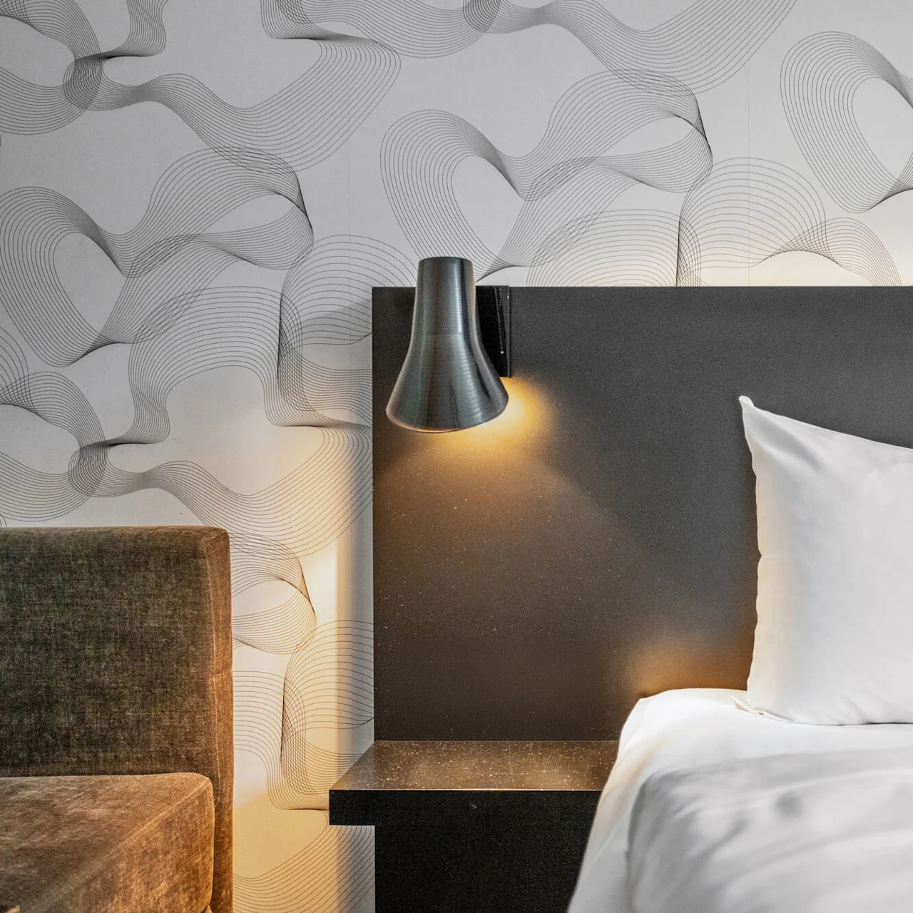 White pillow on a bed with an illuminated wall-mounted lamp; background has a wavy patterned wallpaper.