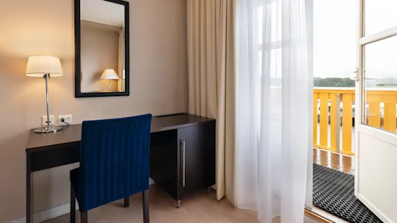 A black desk with a blue chair stands in a room beside a lamp and mirror, next to sheer curtains revealing an open door leading to a balcony with yellow railing.