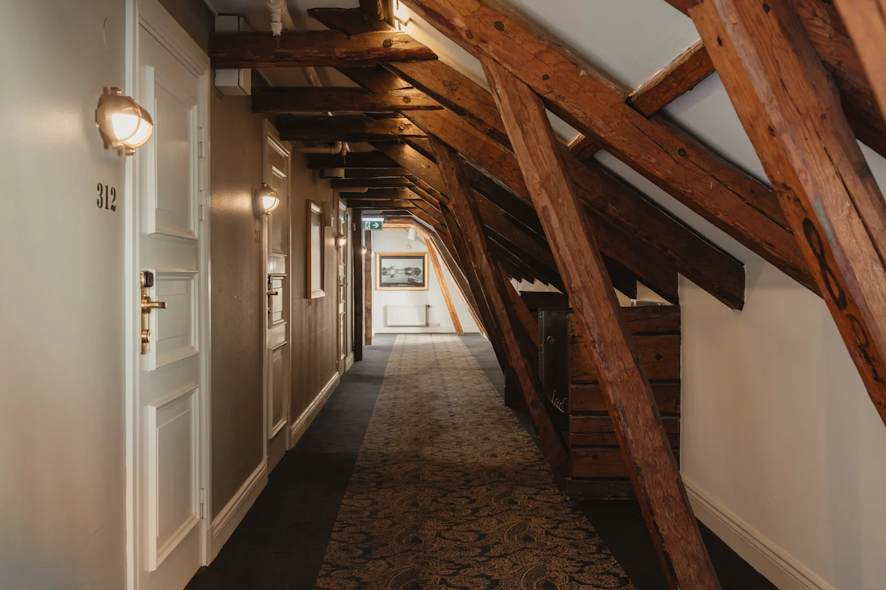 En hyggelig hotelgang med synlige træbjælker i loftet, mønstret tæppe og døre, der fører til værelser. Værelse 312 er synligt.