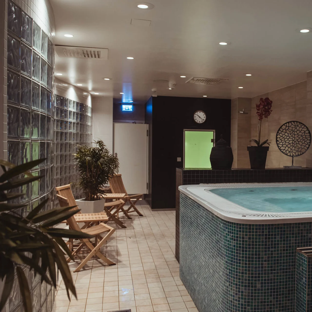 A hot tub sits steaming, surrounded by wooden chairs and potted plants, in a tiled spa room. Glass blocks form a decorative wall. A clock and art decor hang nearby.