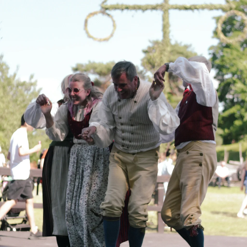People in traditional attire dancing and celebrating midsummer in Sweden, with a maypole in the background.