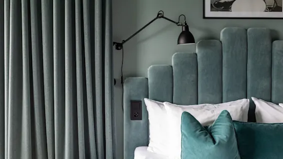 En koselig soveromsscene med en plysjaktig turkis sengegavl, hvite og turkise puter, en veggmontert lampe og matchende turkise gardiner.