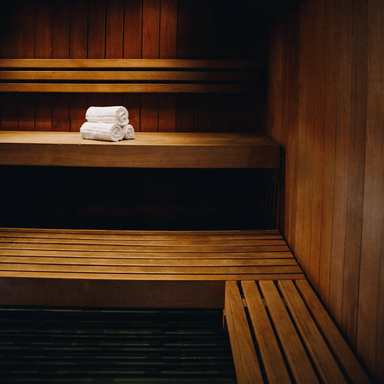 Wooden benches, arranged in a sauna, hold neatly stacked white towels; warm ambient lighting creates a relaxing atmosphere.