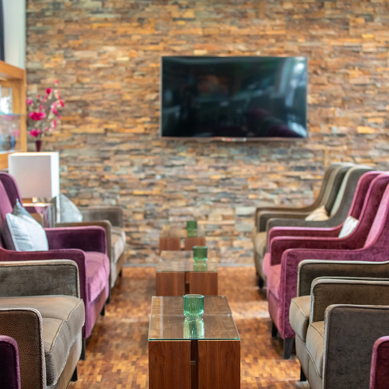 Elegante stoler står overfor hverandre over blanke bord med grønne vaser, i en lounge med steinvegg og montert TV.