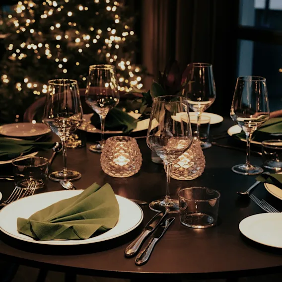 Et elegant dekket rundt middagsbord med vinglass, grønne servietter, levende lys og uskarpe juletrelys i bakgrunnen.