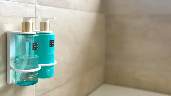 Two bottles of Rituals shower gel and shampoo hang on a tiled bathroom wall, near a bathtub. A stack of folded towels is visible on a ledge in soft focus.