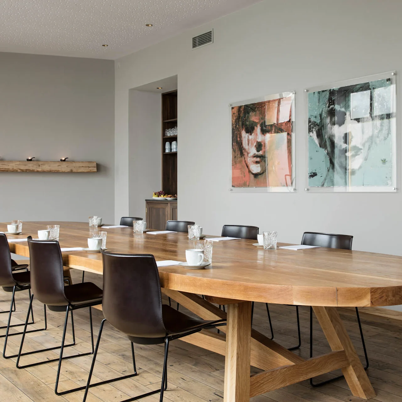 Large wooden table surrounded by chairs, set with cups and glasses, in a conference room. Walls feature modern art and a shelf with a vase above. Wooden floor.