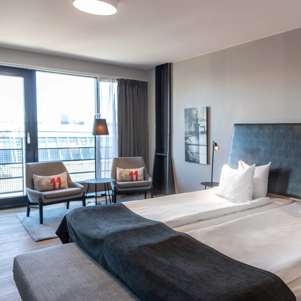 A bed neatly made with white pillows and dark covers stands in a modern hotel room. Two chairs with cushions labeled "11" face a window overlooking buildings.