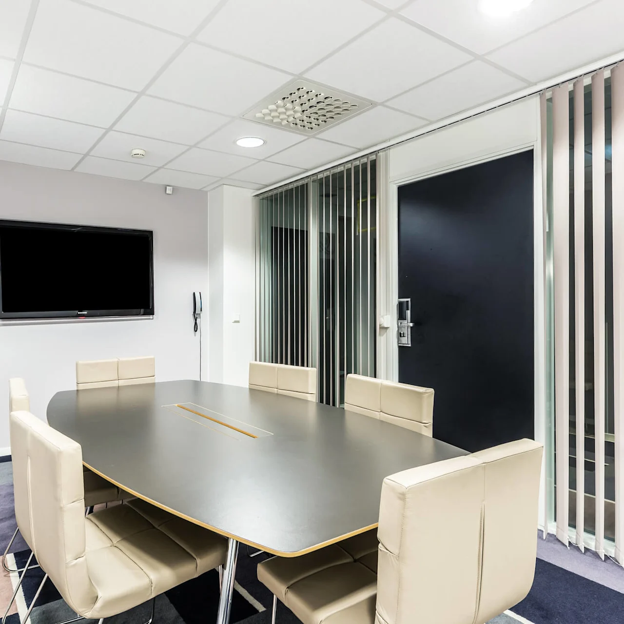 Konferansebord omgitt av beige stoler i et moderne møterom med veggmontert skjerm, vertikale persienner og mønstret teppe. Ingen tekst er til stede.