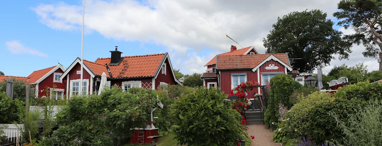 Røde træhuse med gavlede tage står blandt frodige haver med blomstrende roser og velplejede buske. Et svensk flag vifter på en stang under en delvis overskyet himmel.