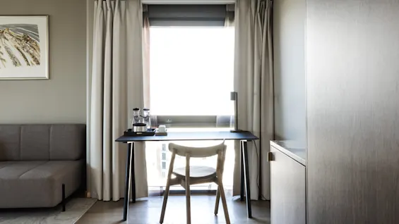 A chair faces a desk set with a coffee tray and lamp by a window with curtains. A gray sofa and wall art are nearby, within a minimalist room.
