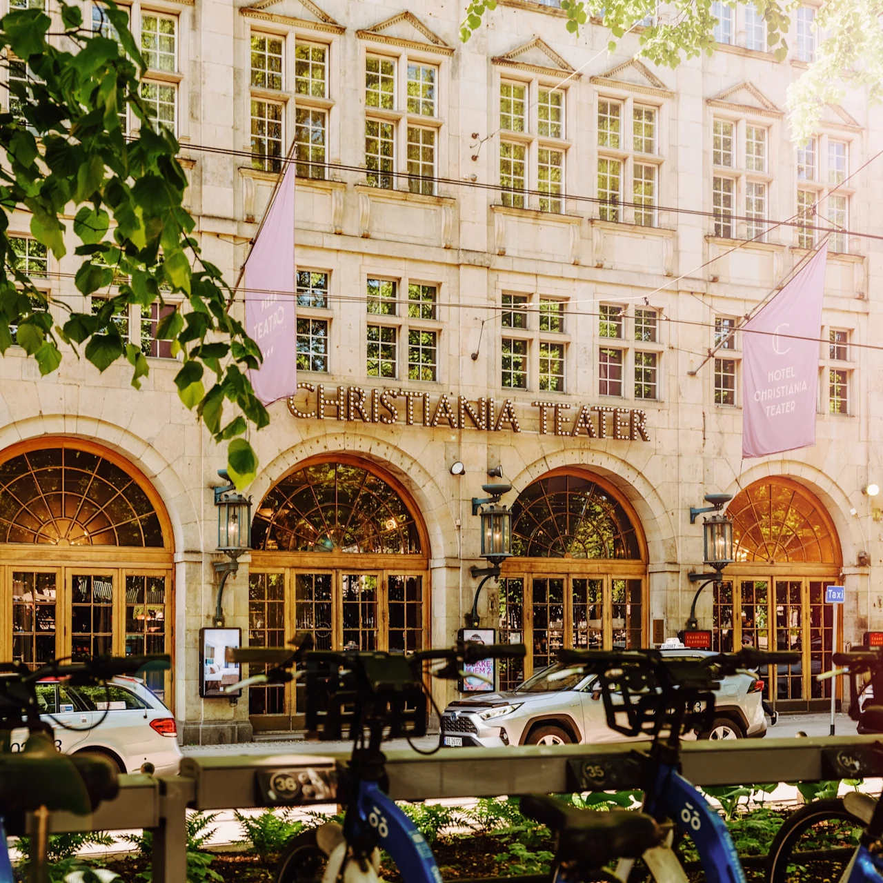 Facade of a historic theater with arched windows, titled "CHRISTIANIA TEATER." Bicycles are parked in the foreground, while cars are visible on the street. Green foliage frames the scene. 