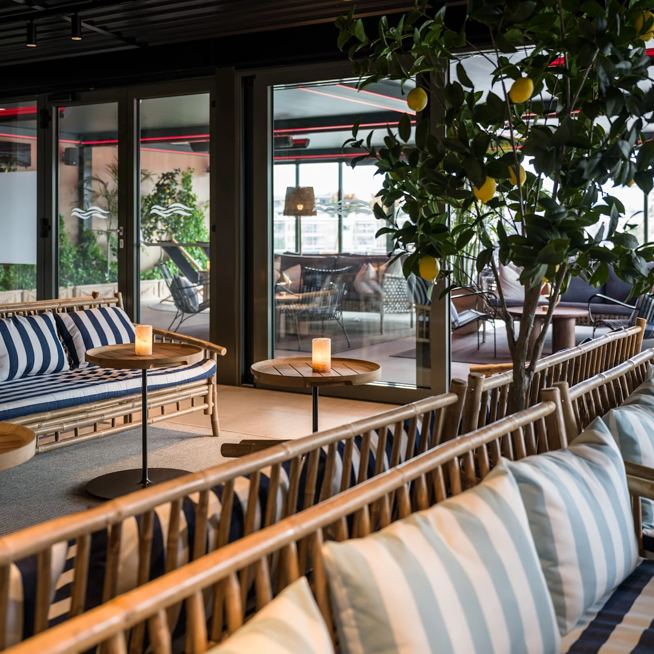 Klädda soffor i trä med blå randiga kuddar står i en lounge bredvid runda bord, med ett krukträd och stora fönster mot en terrass.