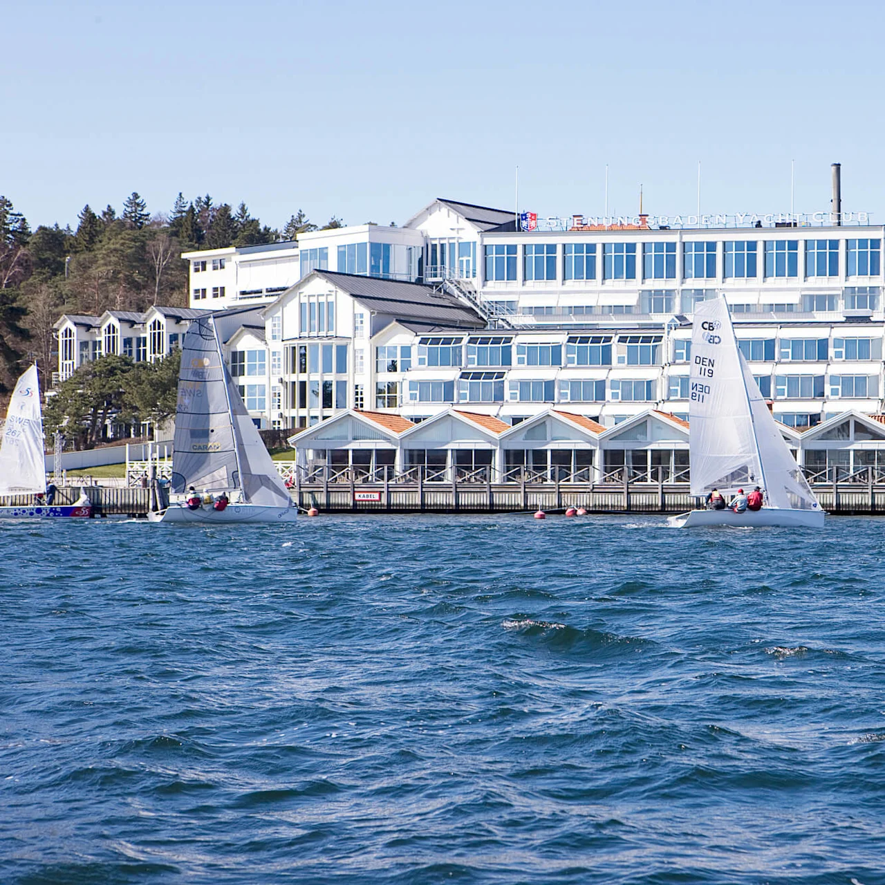 Sailboats glide across a sunny, windy harbor in front of a white, multi-story building labeled "Svendborg Sunds Sejlklub." Surrounding trees create a serene waterfront setting.