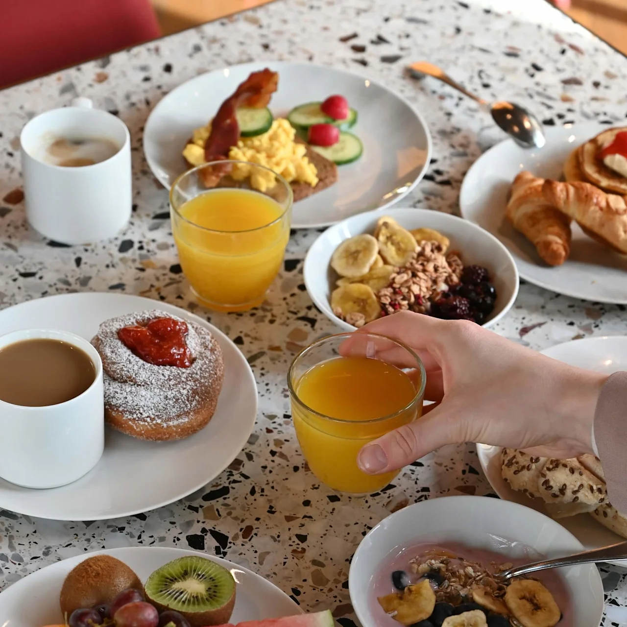 A hand holds a glass of orange juice over a breakfast spread with croissants, fruit, cereal bowls, coffee, and pastries on a speckled countertop.
