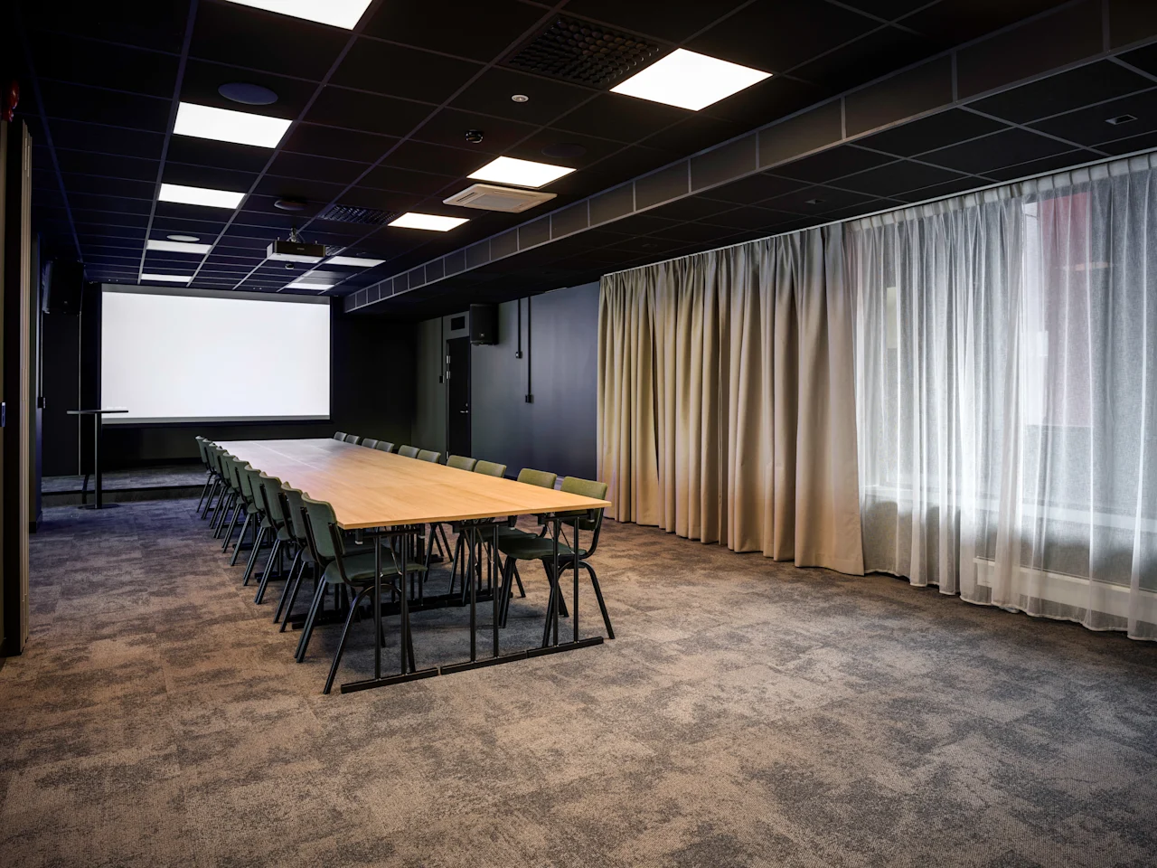 A modern conference room with a long table, chairs, a large projector screen, and sheer curtains, ready for a presentation or meeting.
