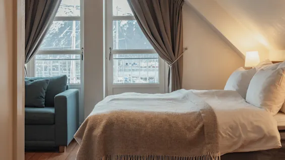 A cozy bedroom with a bed, a sofa, and two windows with curtains. A lamp illuminates the sloped ceiling, creating a warm atmosphere.