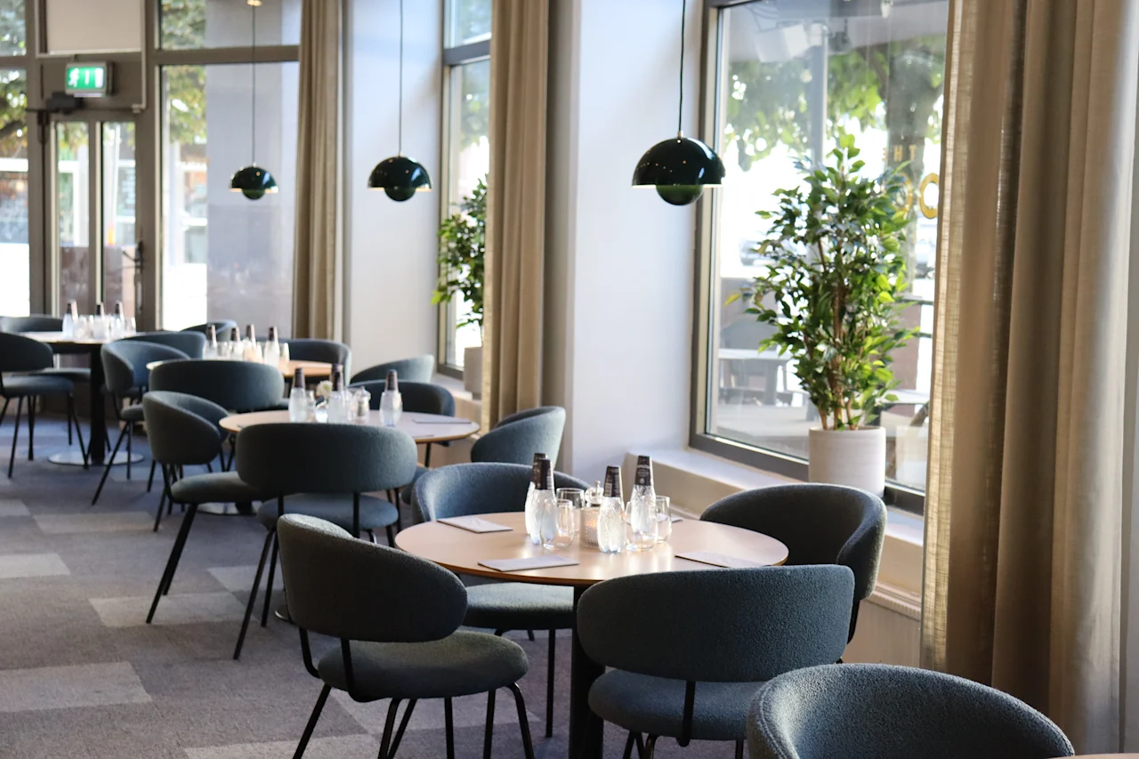 Round tables with chairs are neatly arranged in a modern dining area, featuring large windows and potted plants, creating a bright and inviting ambiance.