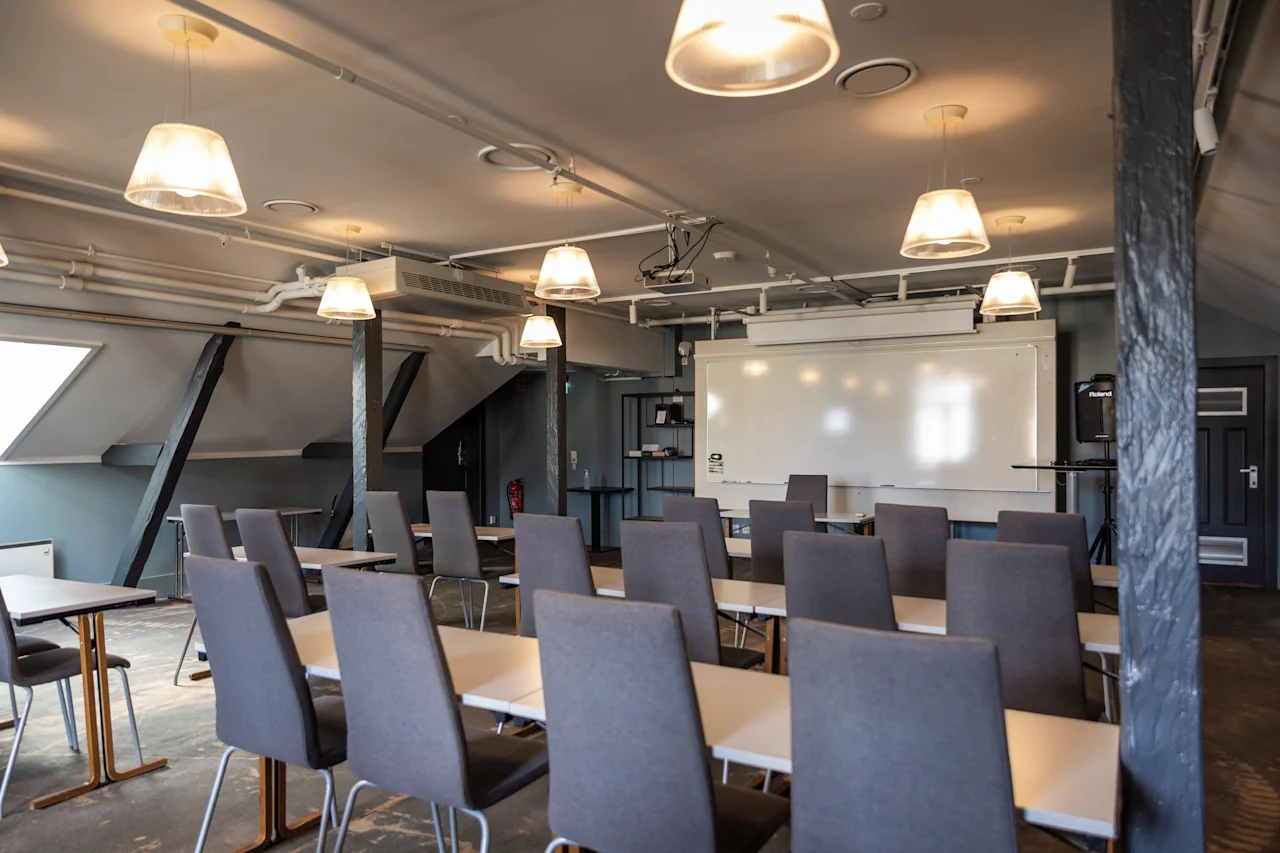 Klassrumsmiljö: Tomma grå stolar är vända mot en whiteboard i ett vinden-stil rum, upplysta av taklampor. Bord är arrangerade för presentation eller föreläsning.