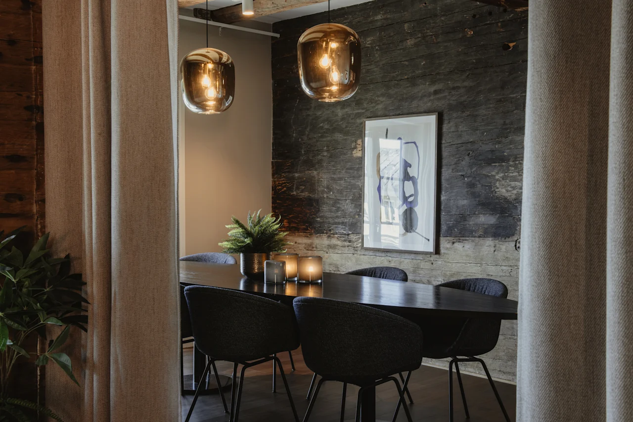 Et stilfuldt interiør med et mørkt bord, moderne stole, varme pendellamper og en rustik trævæg, der skaber en indbydende atmosfære.