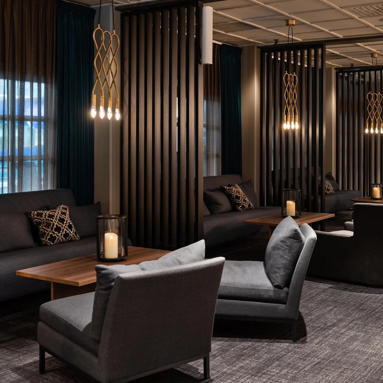 Dark gray sofas and chairs surround wooden tables with candles, under intricate gold light fixtures, in a cozy lounge featuring wooden dividers and curtained windows.