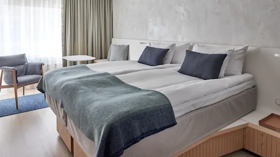 A neatly made double bed with blue and gray pillows and blanket stands in a modern, bright room. A gray armchair and white table are near a window with curtains.