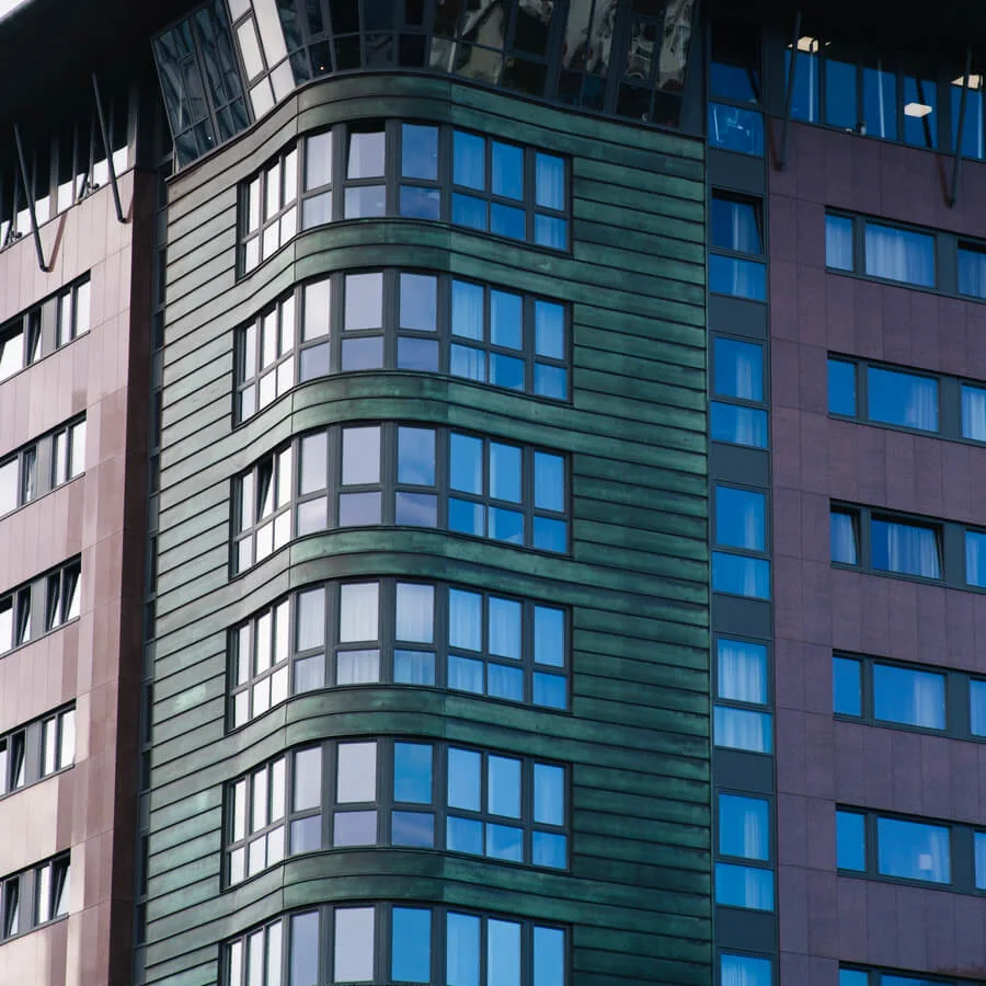 A modern high-rise building features green and brown cladding with large windows. The structure curves slightly outward, located against a partly cloudy sky.
