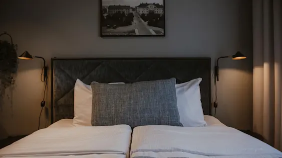 Ett bekvämt hotellrum med en prydligt bäddad säng, mörk sänggavel och varm belysning från väggmonterade lampor. En inramad bild hänger ovanför.