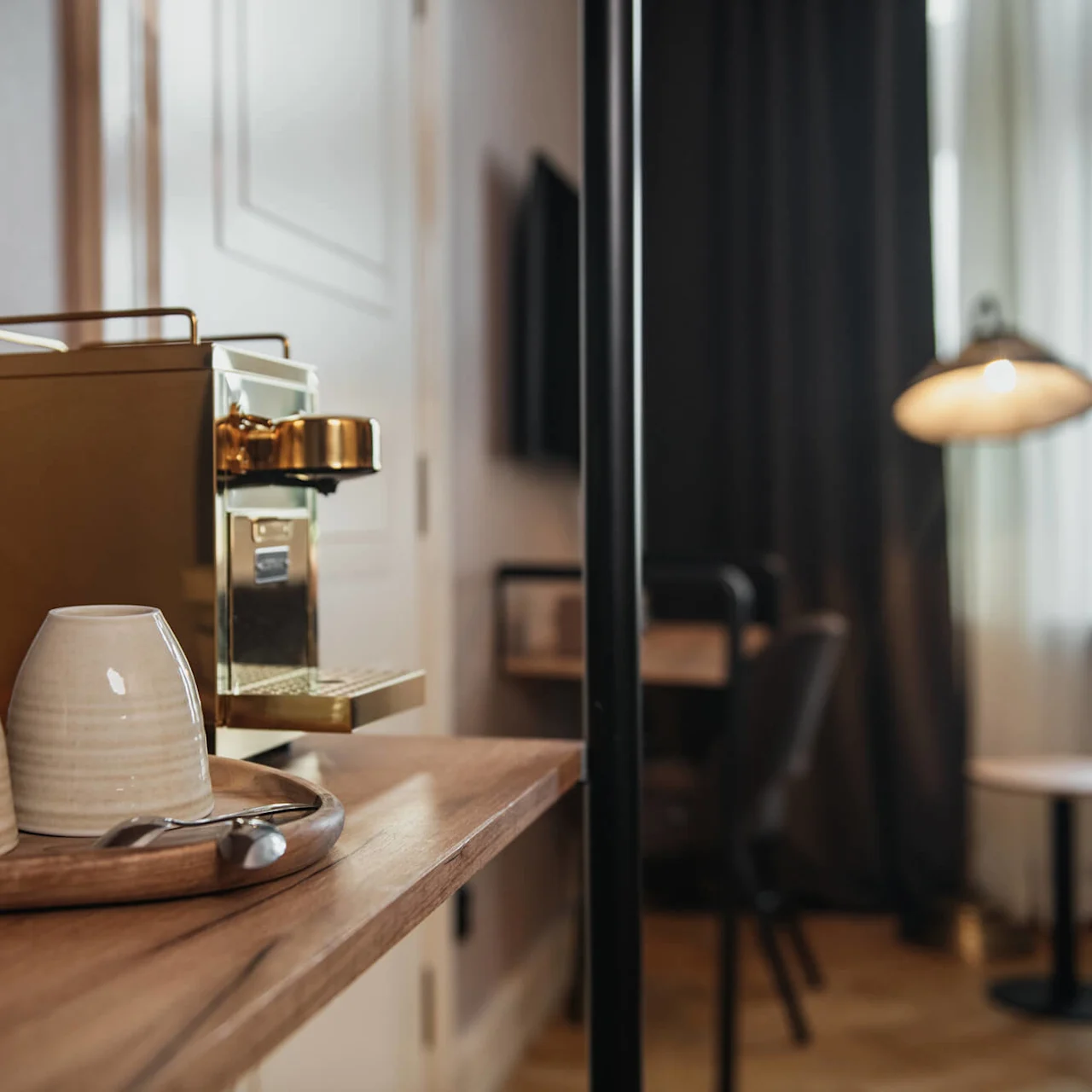 Coffee machine sits on a wooden table near two upside-down ceramic mugs on a tray; a spoon rests nearby. Soft lighting and a blurred room are in the background.