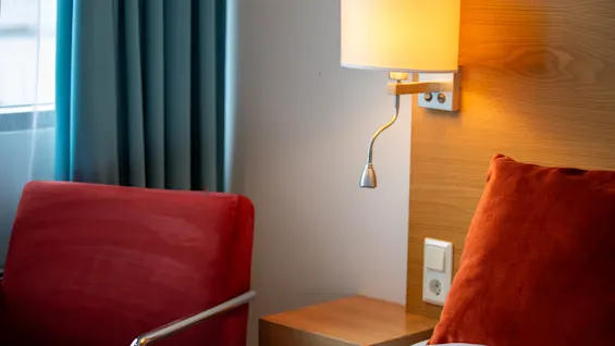 En vegglampe med oransje skjerm er montert på en trepanel, og belyser en rød stol og pute i et rom med turkise gardiner.