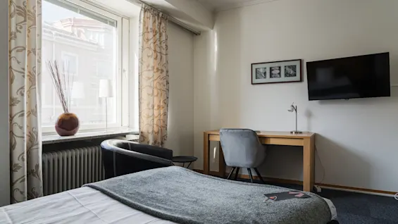 Ett prydligt inrett hotellrum med säng, skrivbord och stol. Solljus strömmar genom ett fönster med tunna gardiner. En väggmonterad TV och inramade bilder kompletterar den moderna inredningen.