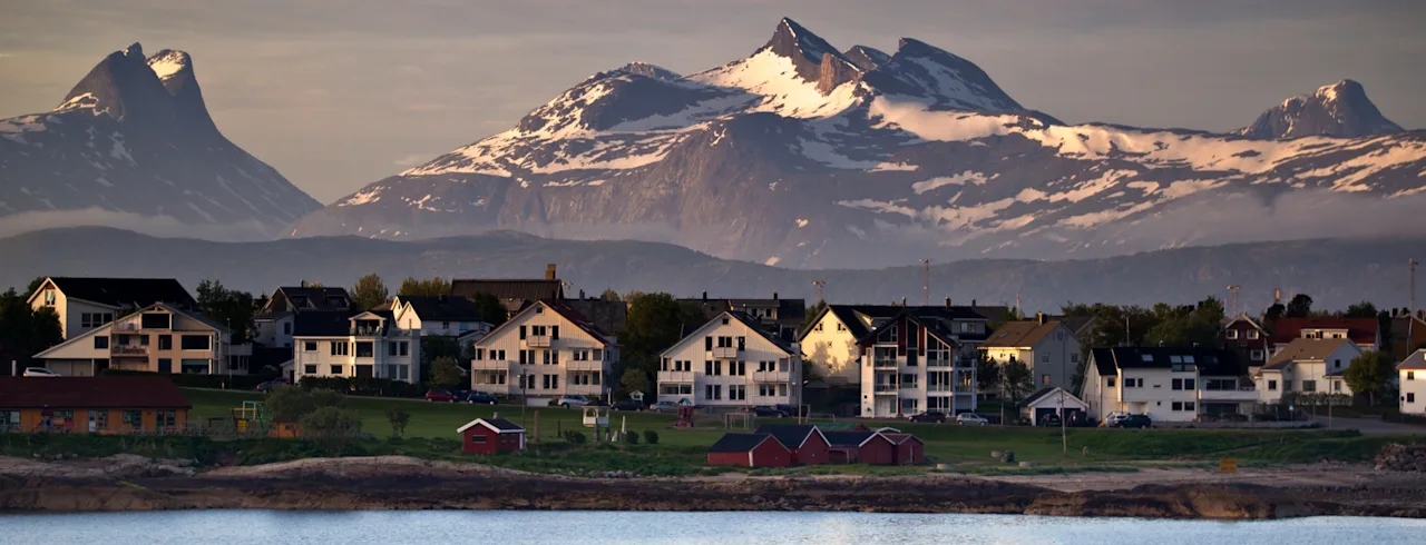 A picturesque coastal town with white houses and red sheds, nestled against a backdrop of majestic, snow-capped mountains.