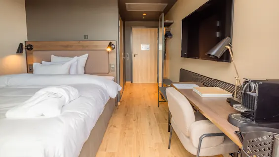 Ett snyggt bäddad säng i ett välbelyst hotellrum med trägolv, ett skrivbord, en stol, en TV och en kaffemaskin.