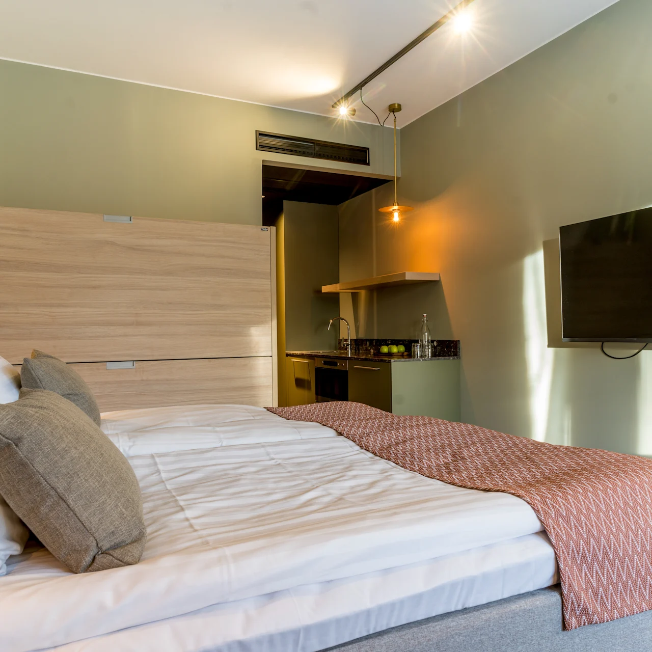 A neatly made bed with gray and white bedding, framed by a wall-mounted TV, in a cozy, modern room with soft lighting, green walls, and a kitchenette in the background.